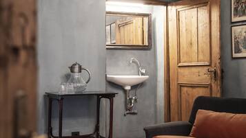 A room with a wooden door, a black sofa, a small table with a glass pitcher, a white sink, and a mirror on the wall.