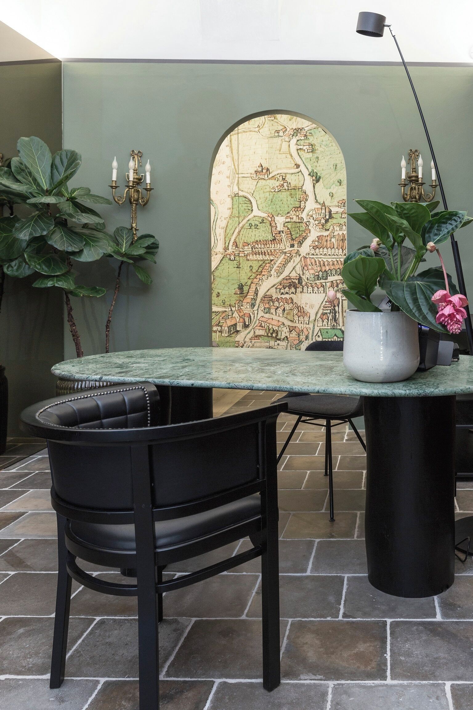 A table with a map mural, a potted plant, and a black chair in a room with stone flooring.