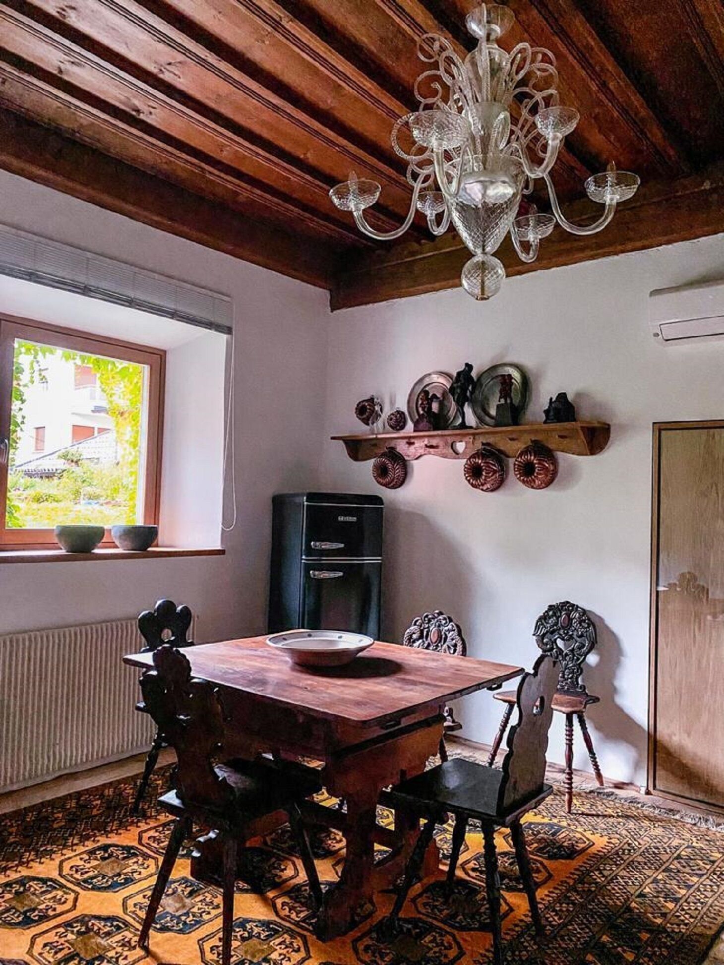 A dining area with a wooden table and chairs, a chandelier, a window with a view, and a shelf with decorative plates.