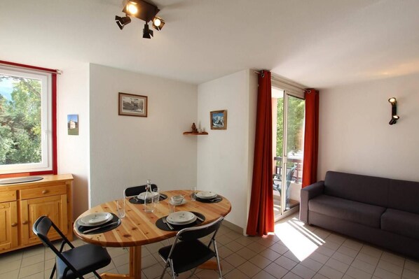 A dining area with a wooden table, chairs, and a sofa. There are two windows with curtains, a ceiling light fixture, and a small shelf with items. - 200m from resort center, 3rd floor, view mountain, balcony, tv, 38m², Serre Chevalier (Briançon)