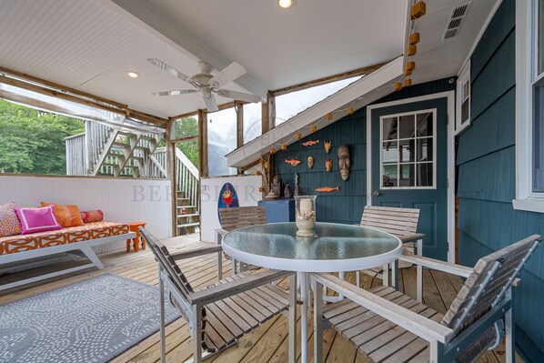 Une terrasse en bois avec une table ronde et des chaises, un canapé agrémenté de coussins colorés et un escalier menant à une autre partie de la terrasse.