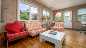 A room with wooden paneling, a red sofa, two floral armchairs, a white coffee table, and large windows with views of greenery.