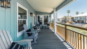 Un porche cubierto con sillas Adirondack, una terraza de madera y vistas a la playa y otros edificios.