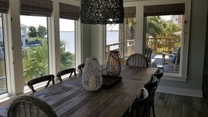 Un comedor con una larga mesa de madera, sillas de mimbre y vistas a un cuerpo de agua y edificios.