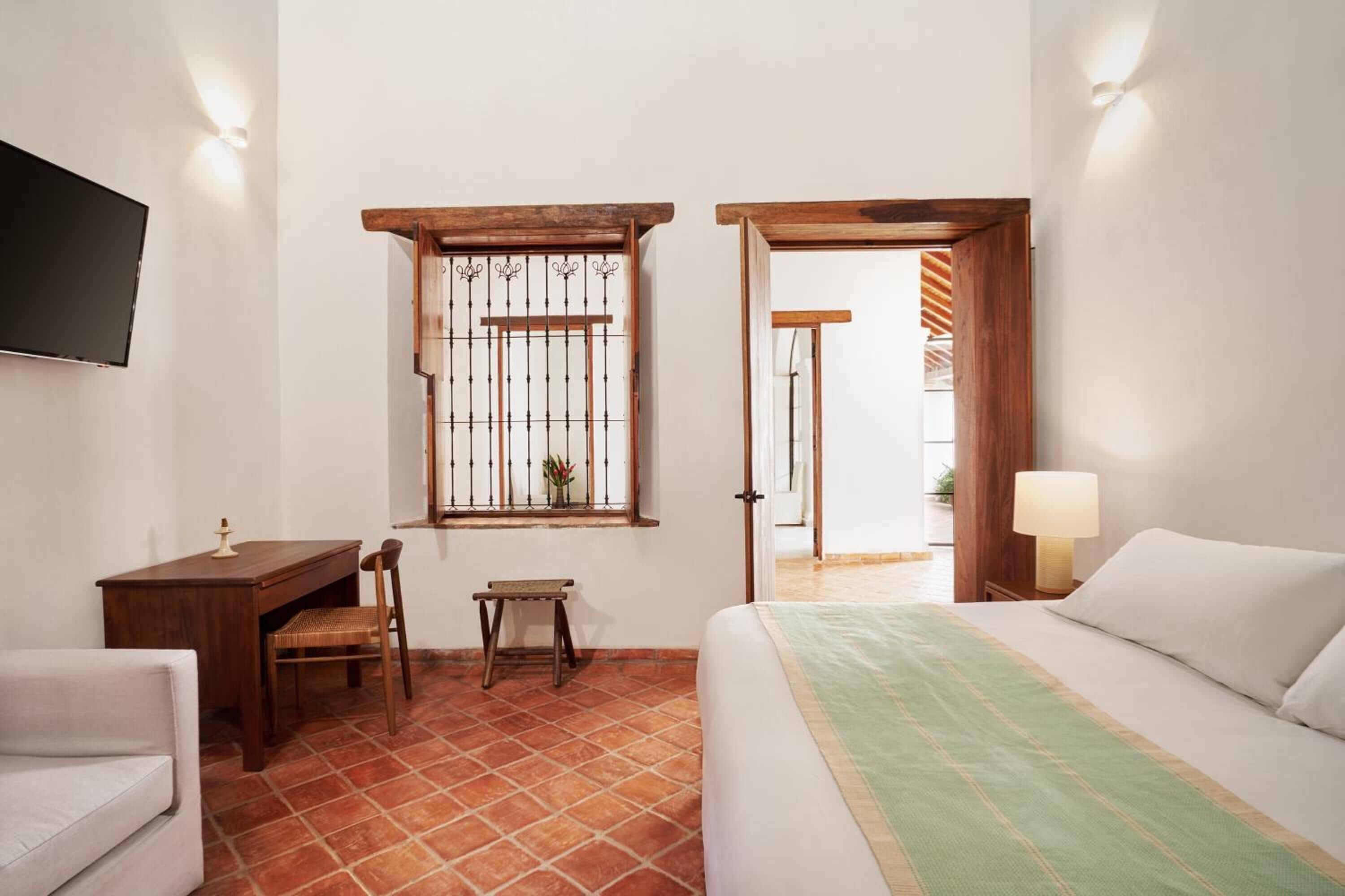 A hotel room with a bed, a desk, a chair, a TV, and a window with a metal grille.