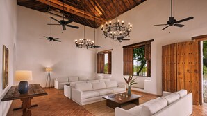 A spacious living room with a white sofa, a coffee table, and a chandelier. There are ceiling fans and a large wooden door.