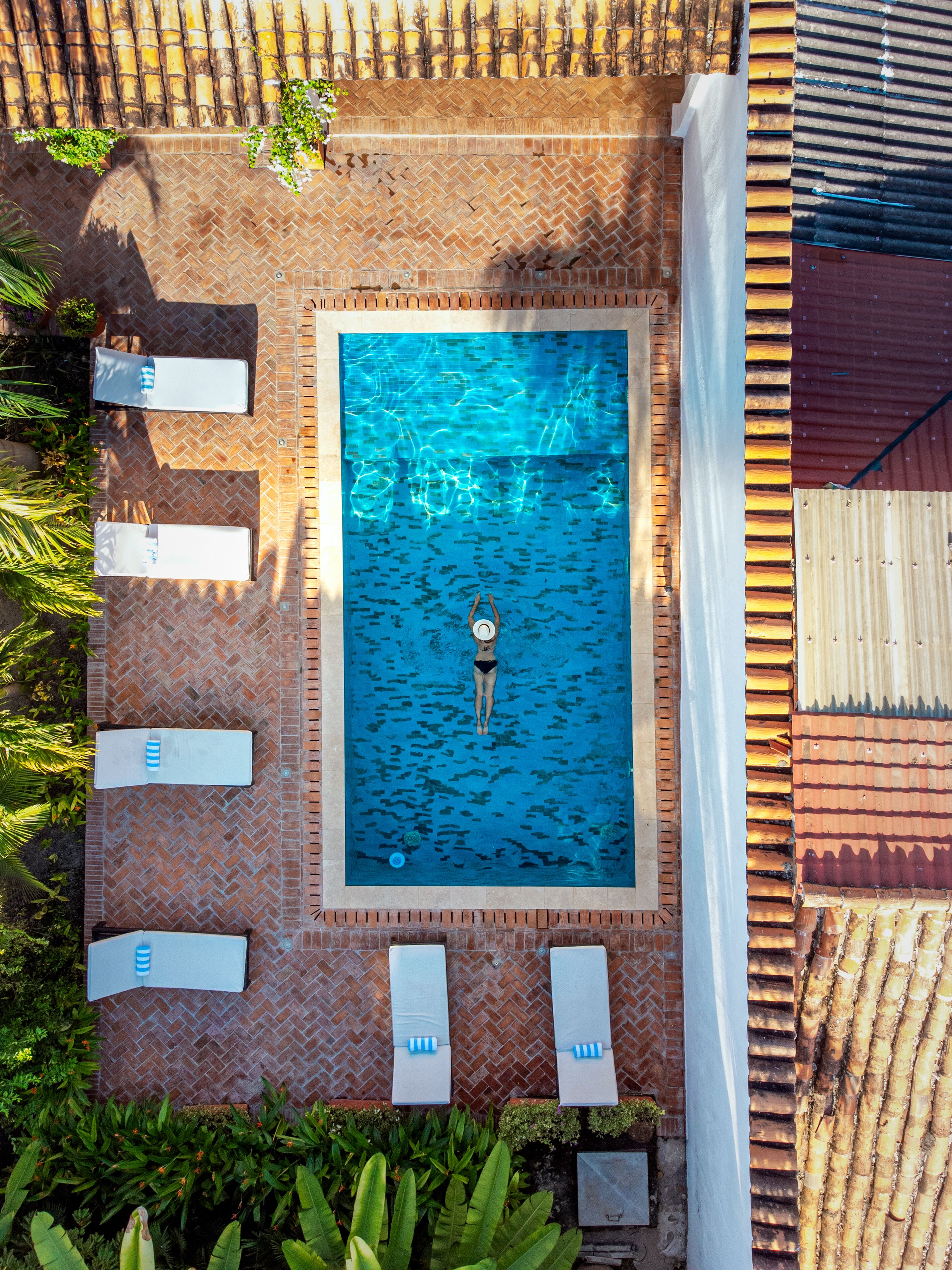 An aerial view of a rectangular swimming pool with a tiled deck surrounding it, equipped with multiple lounge chairs and a person swimming.