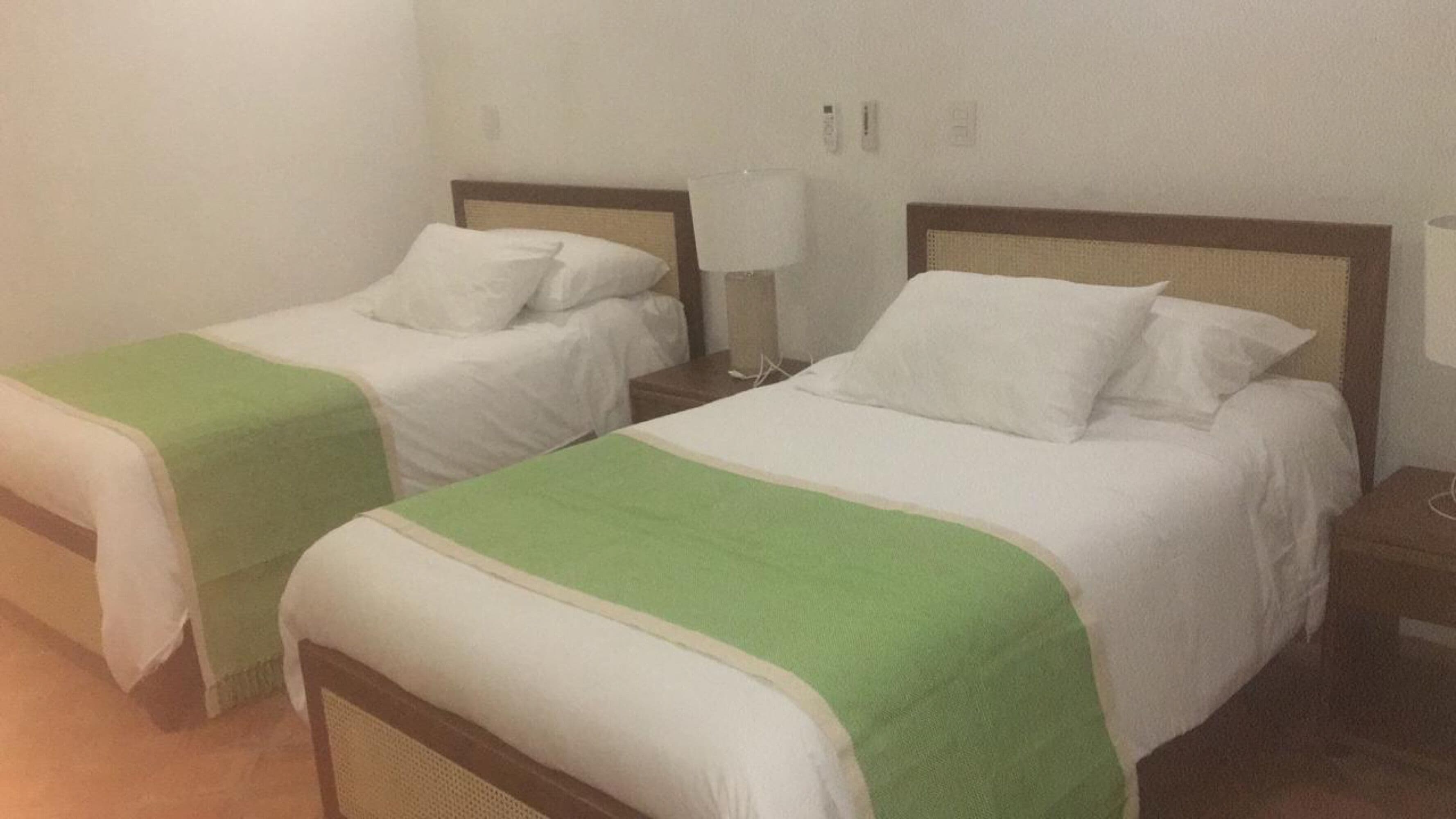 Deux lits simples avec du linge de lit blanc et des plaids verts, chacun étant agrémenté d’une table de chevet et d’une lampe.
