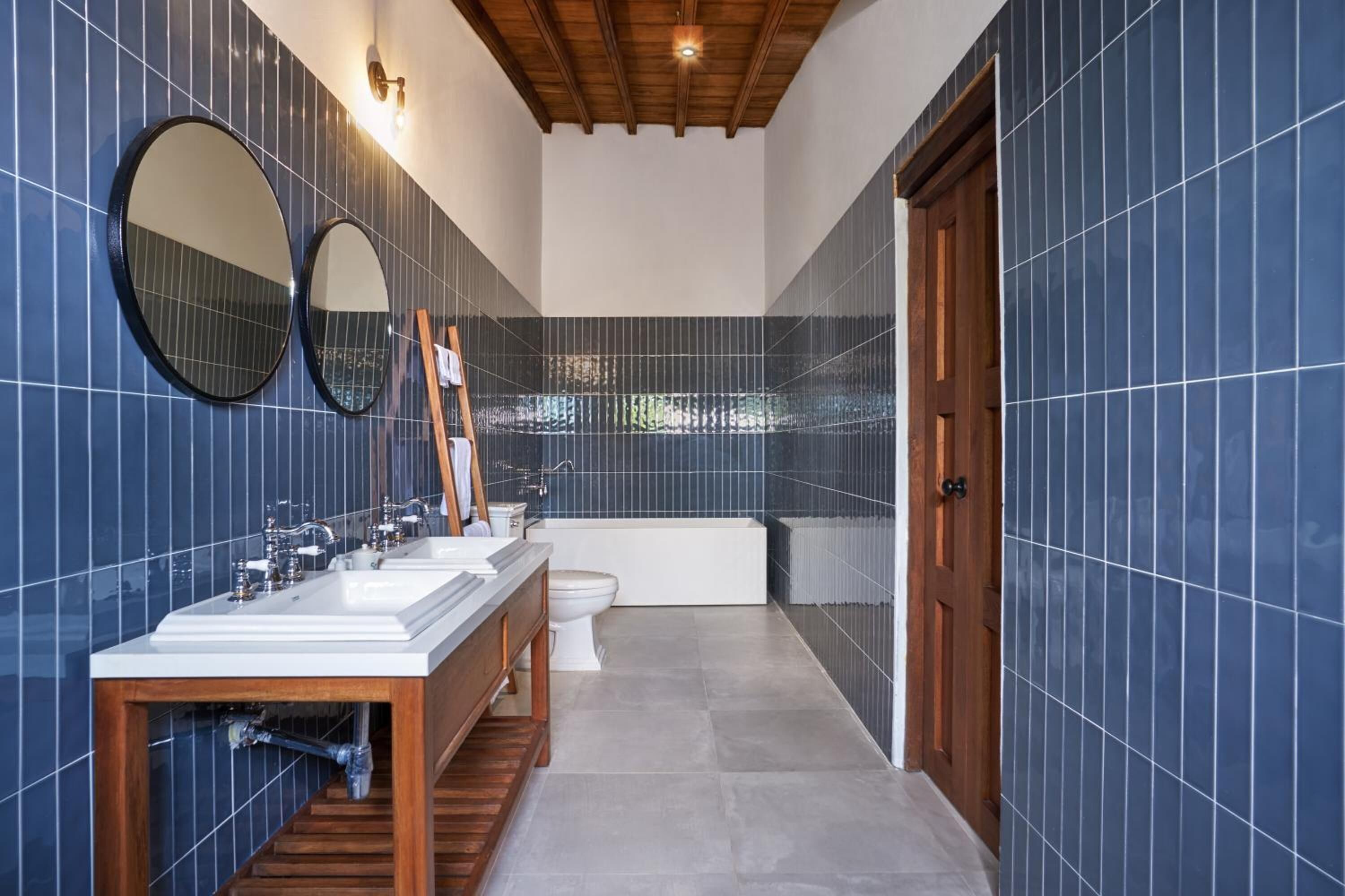 A modern bathroom with blue-tiled walls, a wooden vanity, a bathtub, and two mirrors.