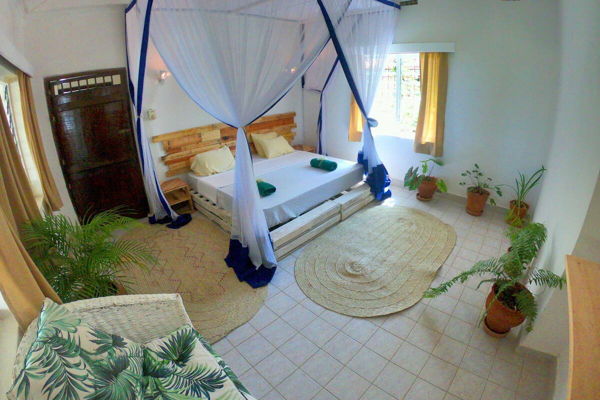 Un dormitorio con cama bajo dosel, cabecera de madera, ventana con cortinas y varias plantas en macetas.