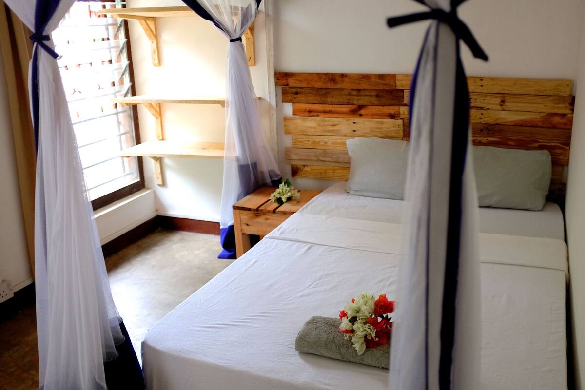 Un dormitorio con cabecera de madera, cama con ropa blanca, mesita de noche y ventana con cortinas translúcidas.