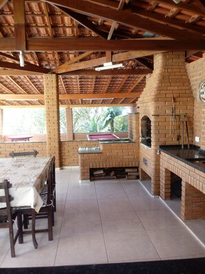 A brick-built outdoor kitchen with a grill, a dining table with chairs, and a window overlooking greenery. - Rent a country house in Ibiúna (Ibiúna)