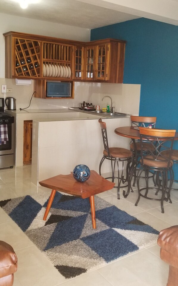 A compact kitchen with wooden cabinets, a microwave, and a sink. There is a dining area with a table and chairs, and a blue and white patterned rug on the floor.