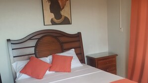 A neatly made bed with white and orange bedding, a wooden headboard, a small bedside table, and a wall-mounted air conditioner. - This property is located near Public  and private beaches and restaurants.. (St Ann)