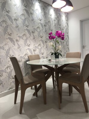 Un área de comedor con una mesa redonda de vidrio rodeada por cuatro sillas, un jarrón con flores y una pared con estampado floral.