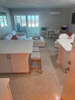 A modern kitchen with a white countertop, a wooden stool, and a dining area with white chairs. - New Listing- Direct Oceanfront -Steps to the SAND -Cocoa Beach Getaway!! (Cocoa Beach)