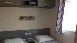 A compact room with two single beds, a small shelf with books, a wall-mounted lamp, and a window with a striped curtain.