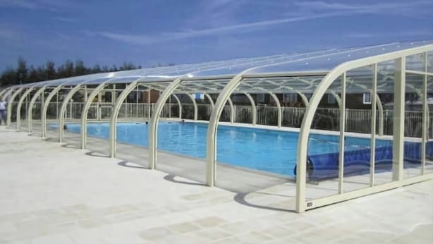 A covered swimming pool with a clear roof and glass walls, surrounded by a tiled deck.