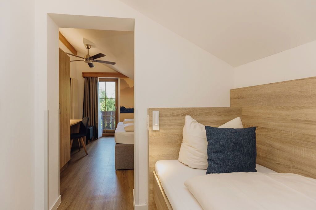 A modern hotel room with a wooden headboard, a bed with white linens and a blue pillow, a ceiling fan, and a view of the outdoors through a large window.