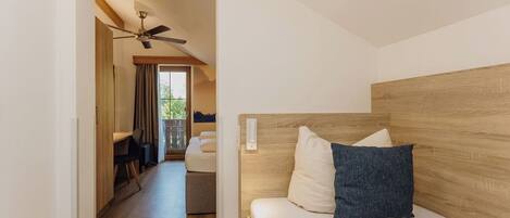 A modern hotel room with a wooden headboard, a bed with white linens and a blue pillow, a ceiling fan, and a view of the outdoors through a large window.