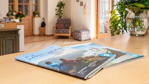 Un magazine est posé sur une table dans une pièce dont le sol est en bois, où se trouvent également une chaise et un vase rempli de fleurs.