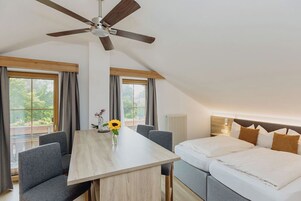 A modern room with a dining table, two gray sofas, a ceiling fan, and a bed with white bedding.