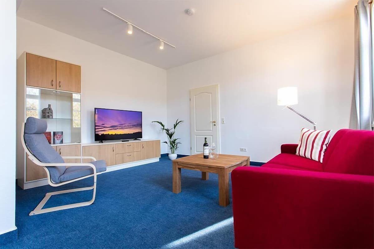 A modern living room with a blue carpet, a red sofa, a wooden coffee table, a TV, and a bookshelf.