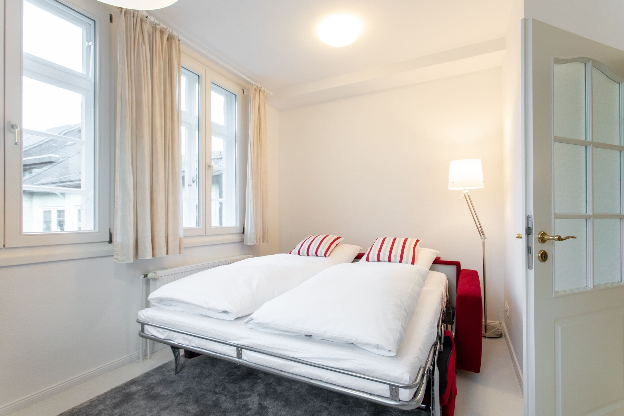 Una habitación de hotel con una cama, una lámpara de noche, una ventana con cortinas y una puerta.