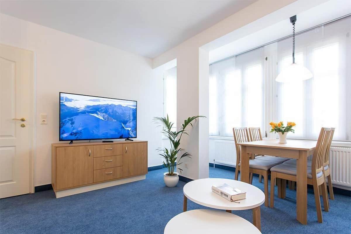 A modern living room with a flat-screen TV, wooden furniture, a potted plant, and a dining area with a table and chairs.