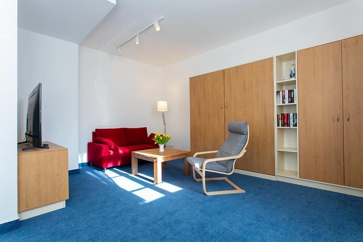 A modern room with a blue carpet, a red sofa, a wooden desk with a TV, a grey chair, and a bookshelf.