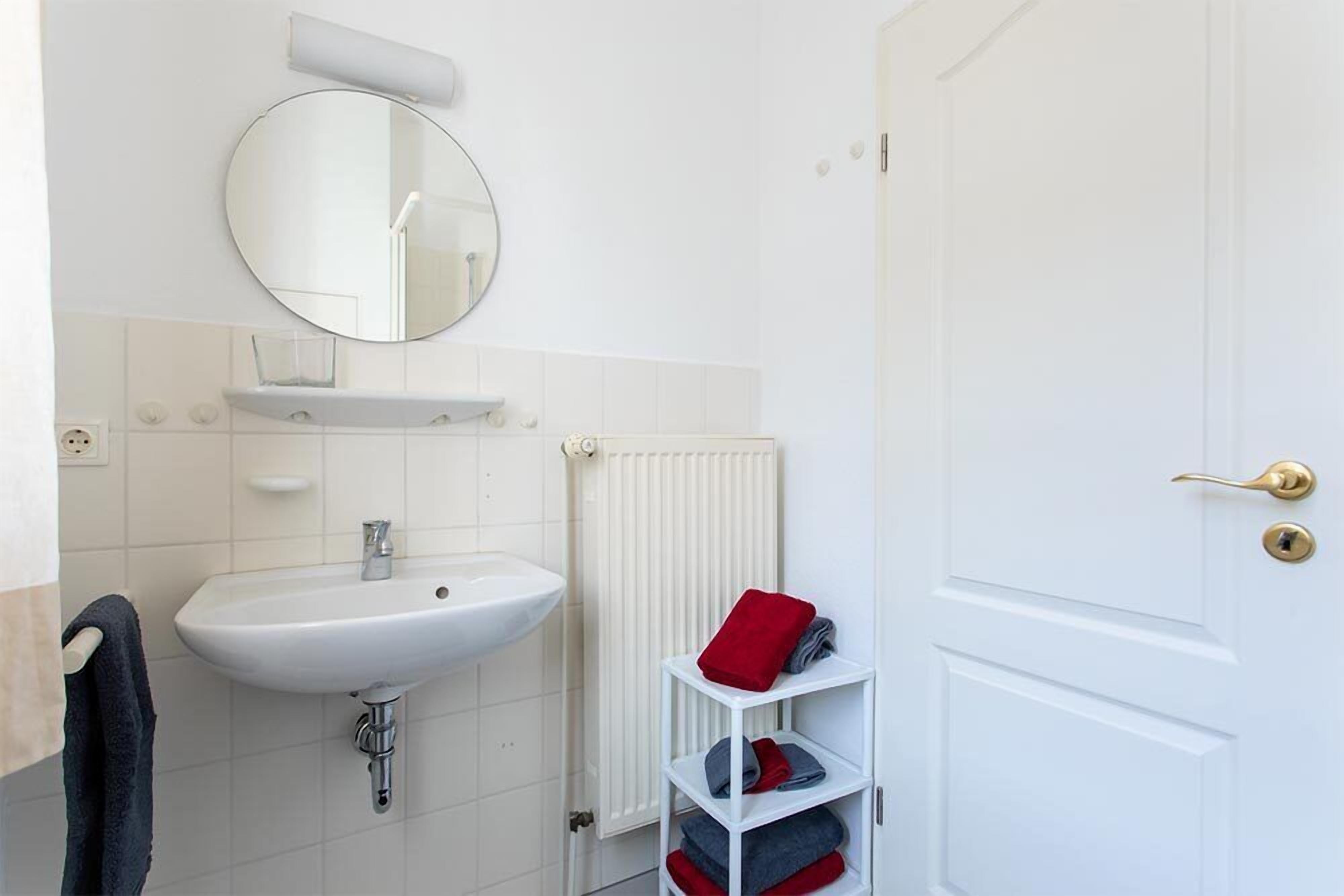 A bathroom with a white sink, a round mirror, a radiator, and a white door with a gold handle.
