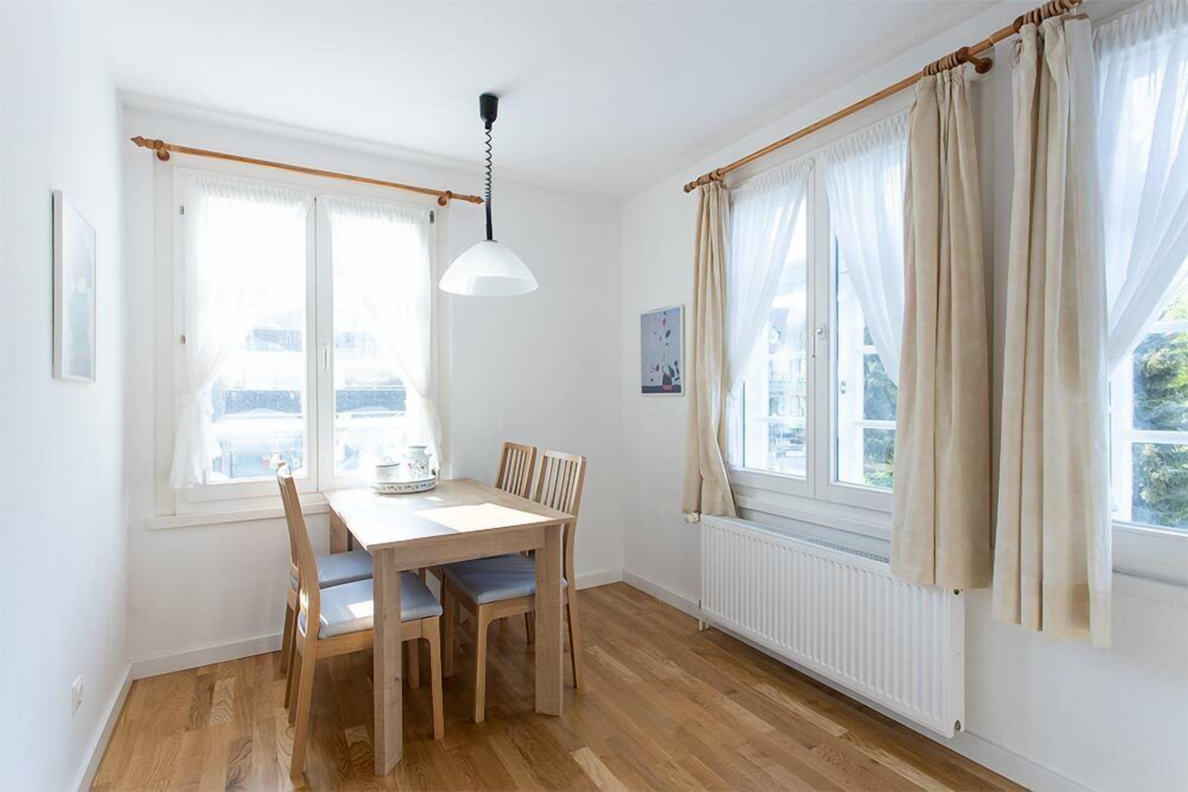 Un comedor con una mesa y sillas de madera, una lámpara colgante y ventanas grandes con cortinas.