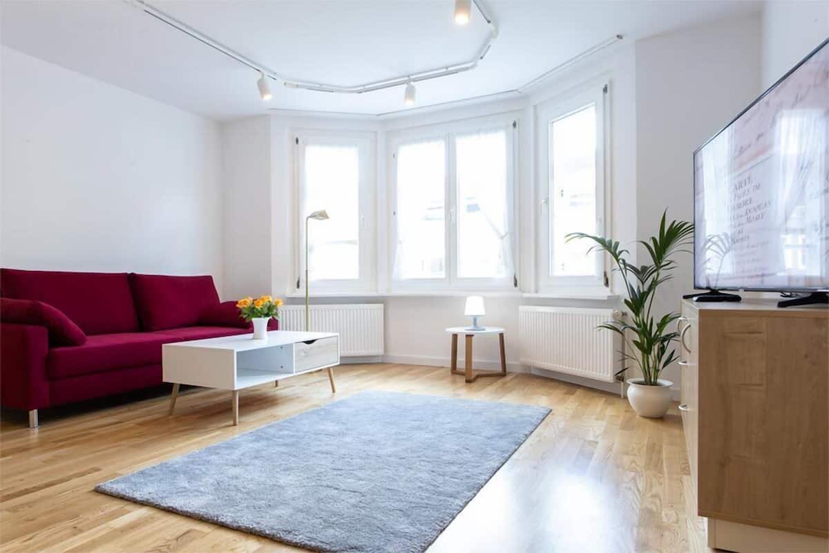 A modern living room with a red sofa, white coffee table, and a grey rug. There are large windows providing natural light, a wooden cabinet, and a potted plant.