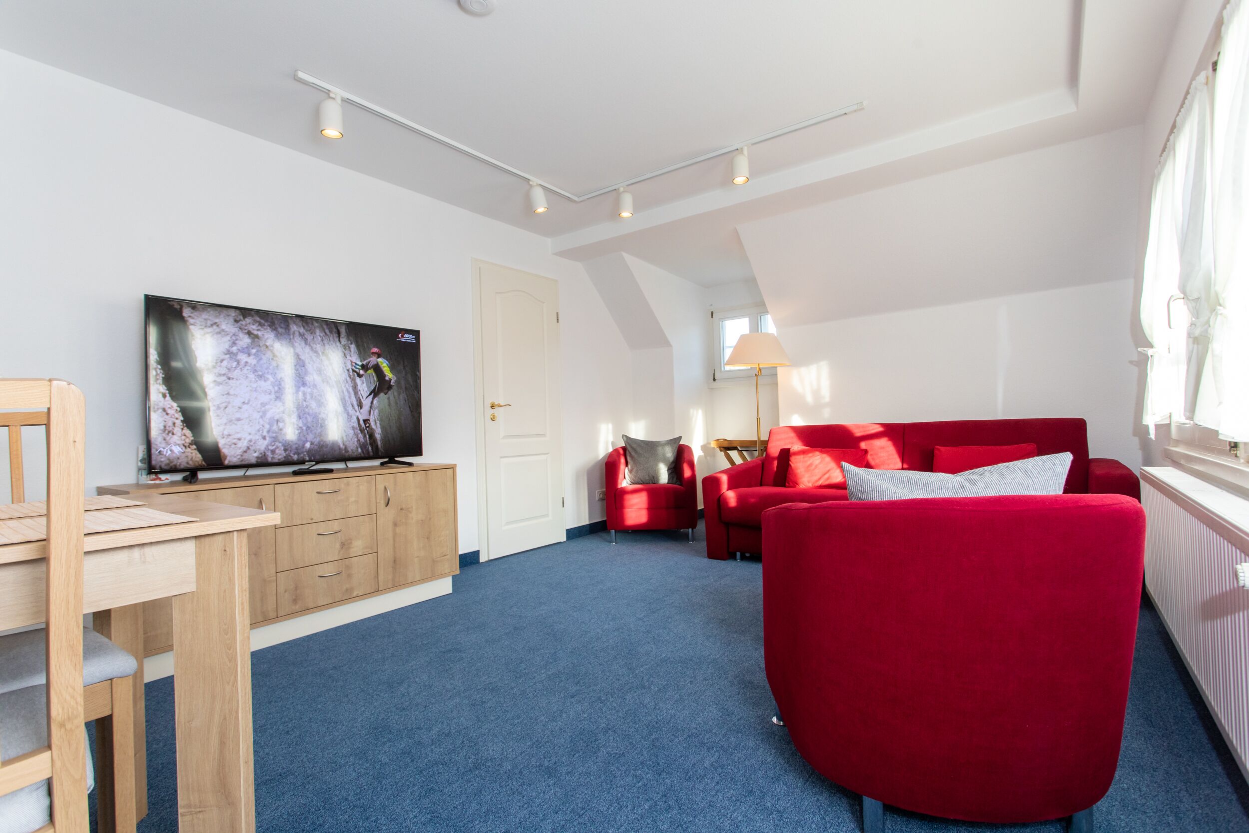 A modern living room with a flat-screen TV, red sofas, a wooden desk, and a blue carpet.