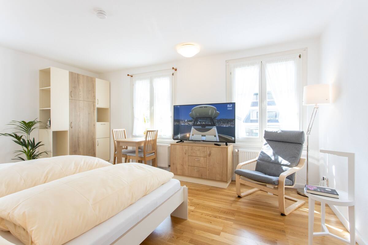 A modern hotel room with a bed, a desk with a TV, a chair, and a plant.