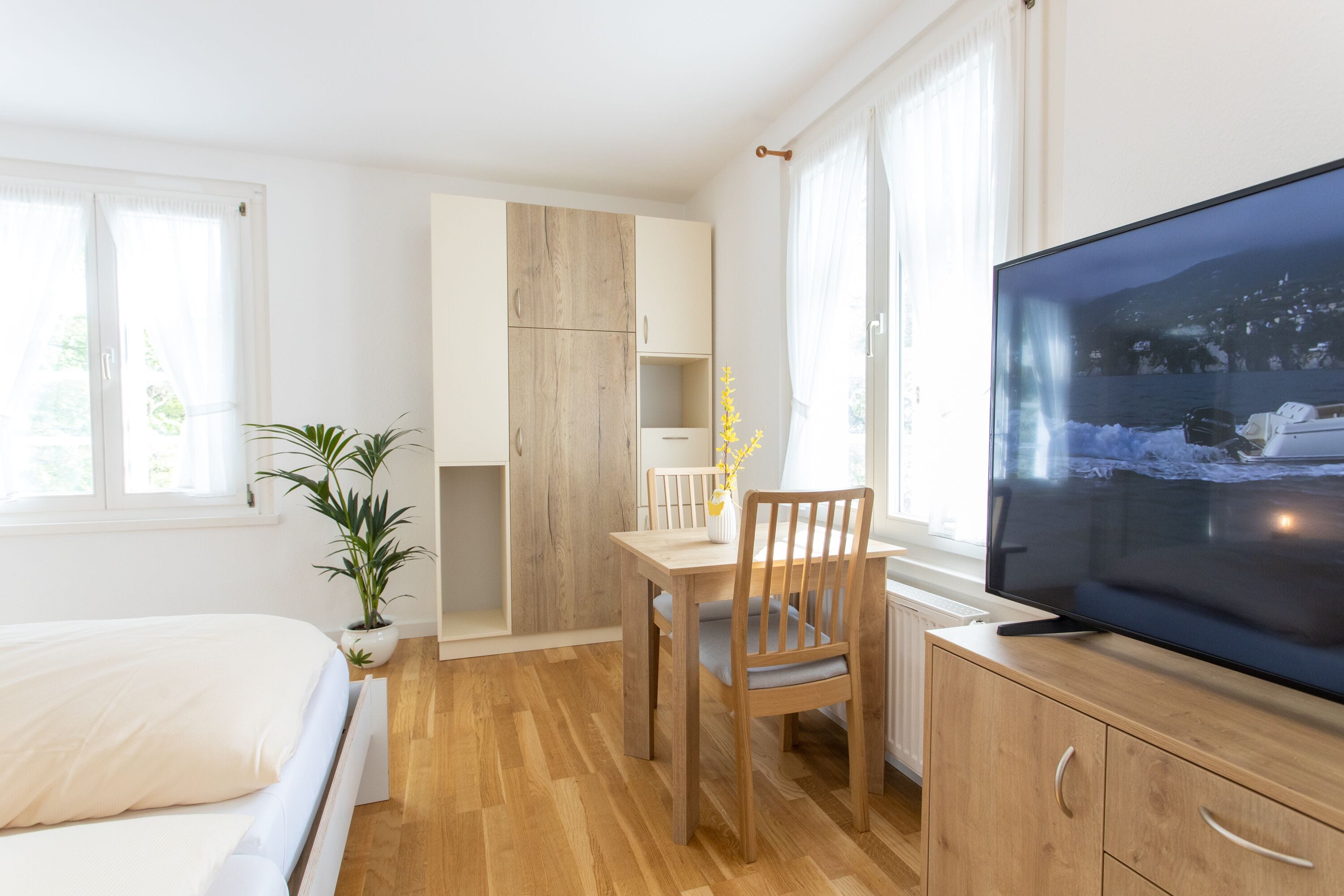 Un dormitorio moderno con una cama, un escritorio de madera, una silla, un televisor y una planta en maceta.