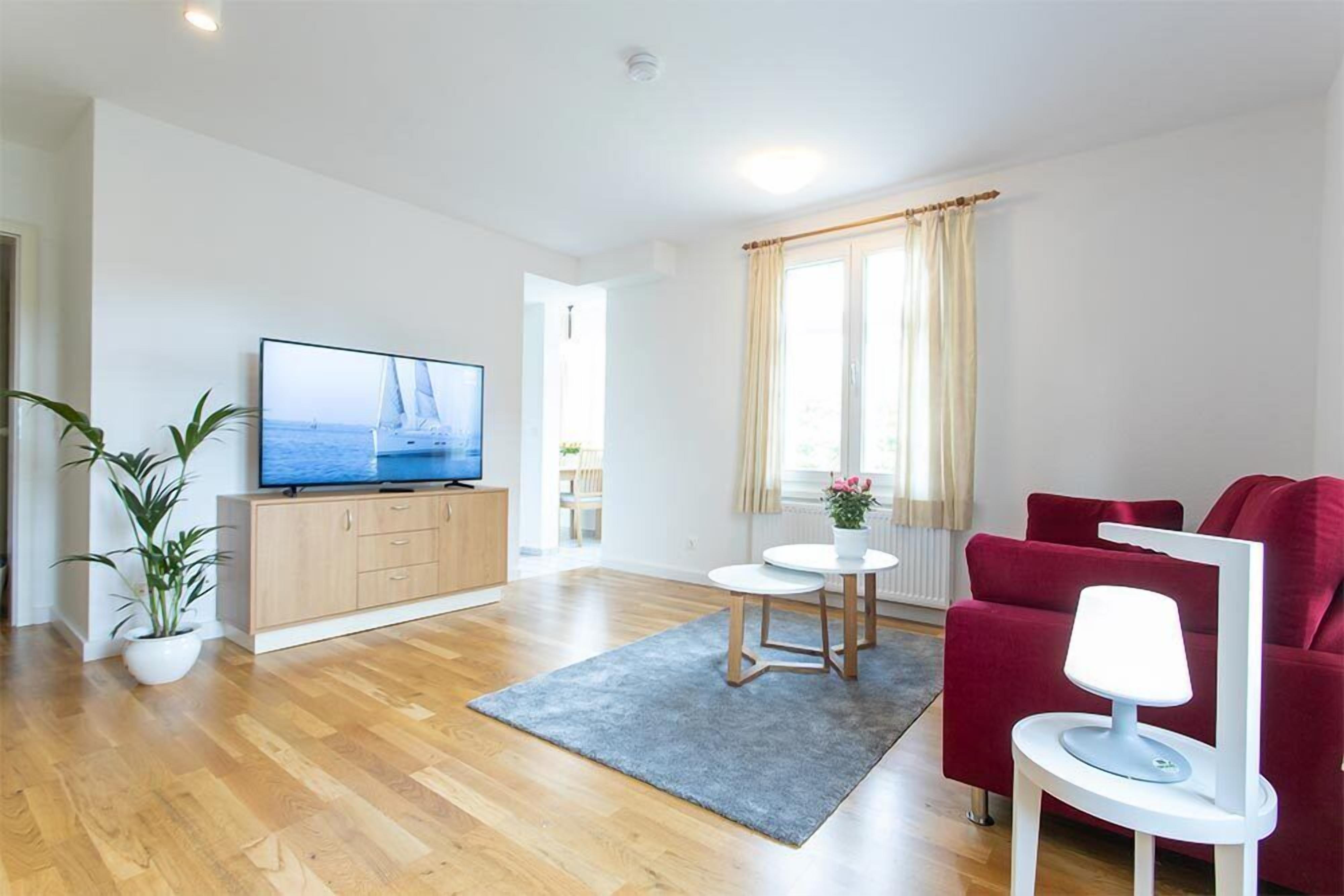 En moderne stue med trægulv, en rød sofa, et fjernsyn på et træskab, et rundt bord med en lampe og en potteplante.