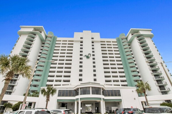 A multi-story hotel with a central entrance, balconies on each floor, and palm trees in the foreground.
