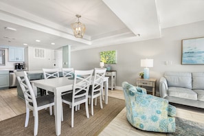 Eine moderne Küche mit weißen Schränken, ein Essbereich mit weißem Tisch und Stühlen, ein graues Sofa und ein blaues, gemustertes Sessel.