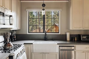 Una cocina con estufa, pileta y refrigerador. Se ve un ventanal con vistas a árboles y una construcción de madera en el exterior.