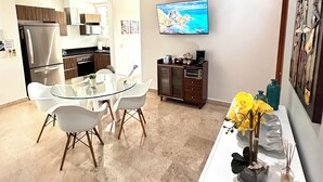 Een moderne keuken met een glazen eettafel, witte stoelen, een aan de muur bevestigde televisie en een decoratieve vaas met gele bloemen.