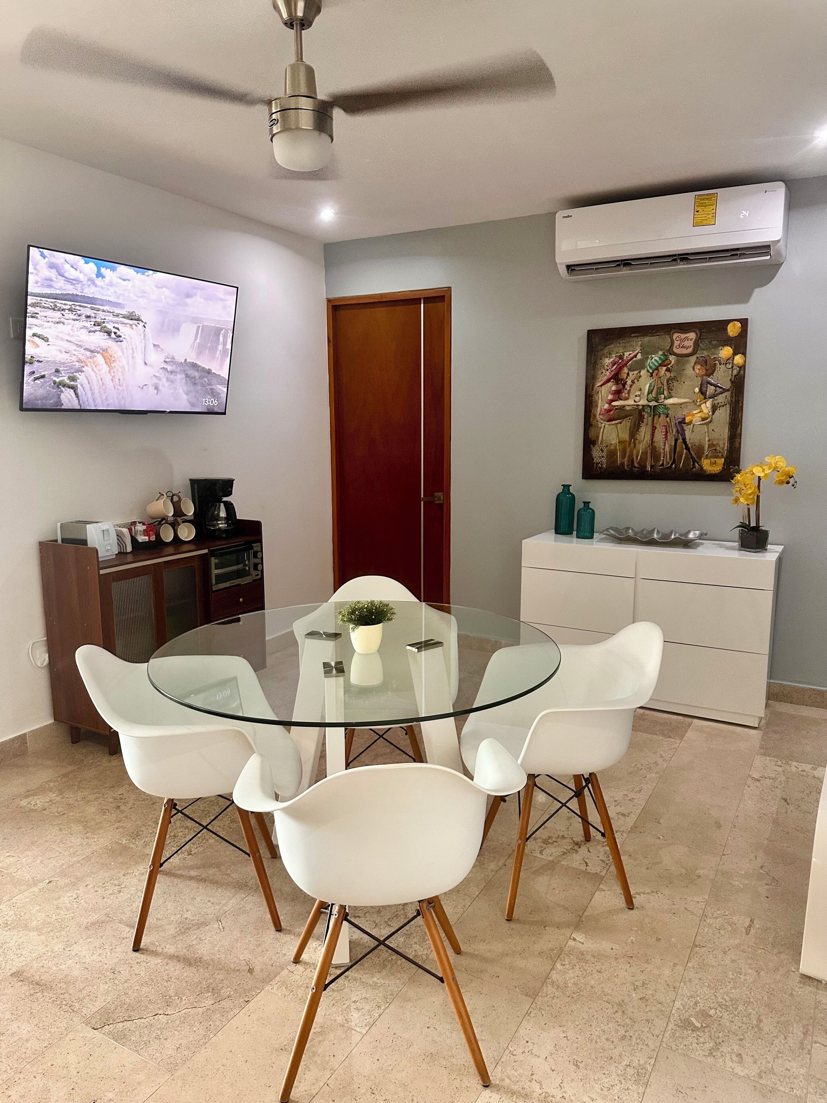 A modern dining area with a glass table, white chairs, a ceiling fan, a TV on the wall, and a painting above a white cabinet.