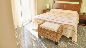 Un lit bien fait, recouvert d’une couvre-lit à rayures, un banc en osier avec un coussin et une table de chevet avec une lampe.