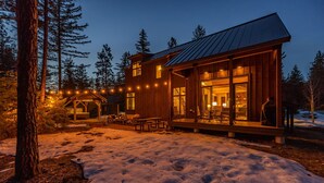 A wooden cabin with large windows and a deck, surrounded by snow and trees.