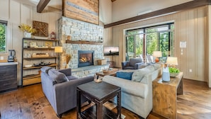 A cozy living room with a stone fireplace, wooden beams, and large windows offering a view of the outdoors.