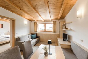 Ein modernes, gut beleuchtetes Zimmer mit Holzbalkendecke, einer Couch, einem Tisch mit einer Vase, einem Fernseher und einem Fenster mit Vorhängen.