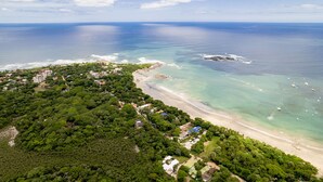 An aerial view of a coastal area with a beach, dense greenery, and scattered buildings.