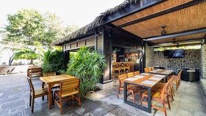 An outdoor seating area with wooden tables and chairs, a stone wall, and a thatched roof.