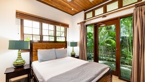 A bedroom with a large bed, two bedside tables, a wooden ceiling, and a view of a tropical garden through a glass door.