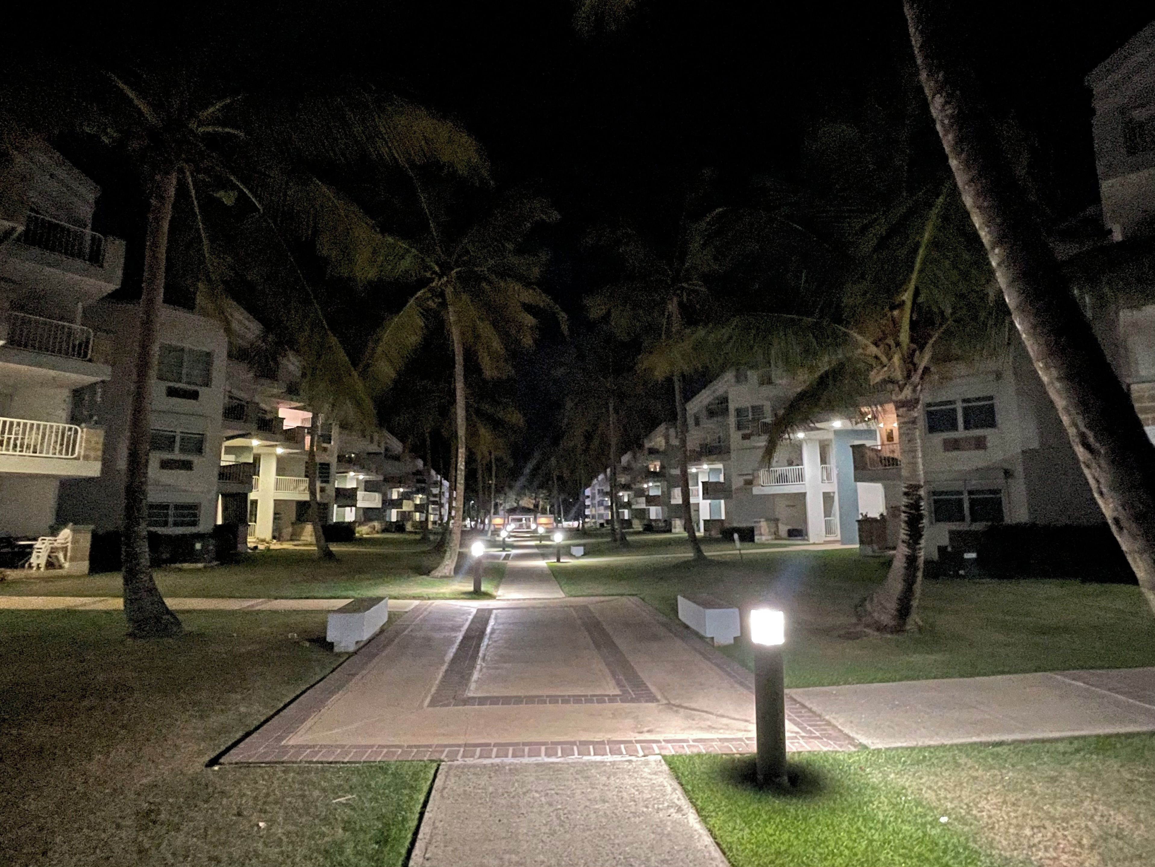 Une vue nocturne d’un complexe hôtelier avec des bâtiments à plusieurs étages, des palmiers et un chemin bien éclairé menant à l’entrée.